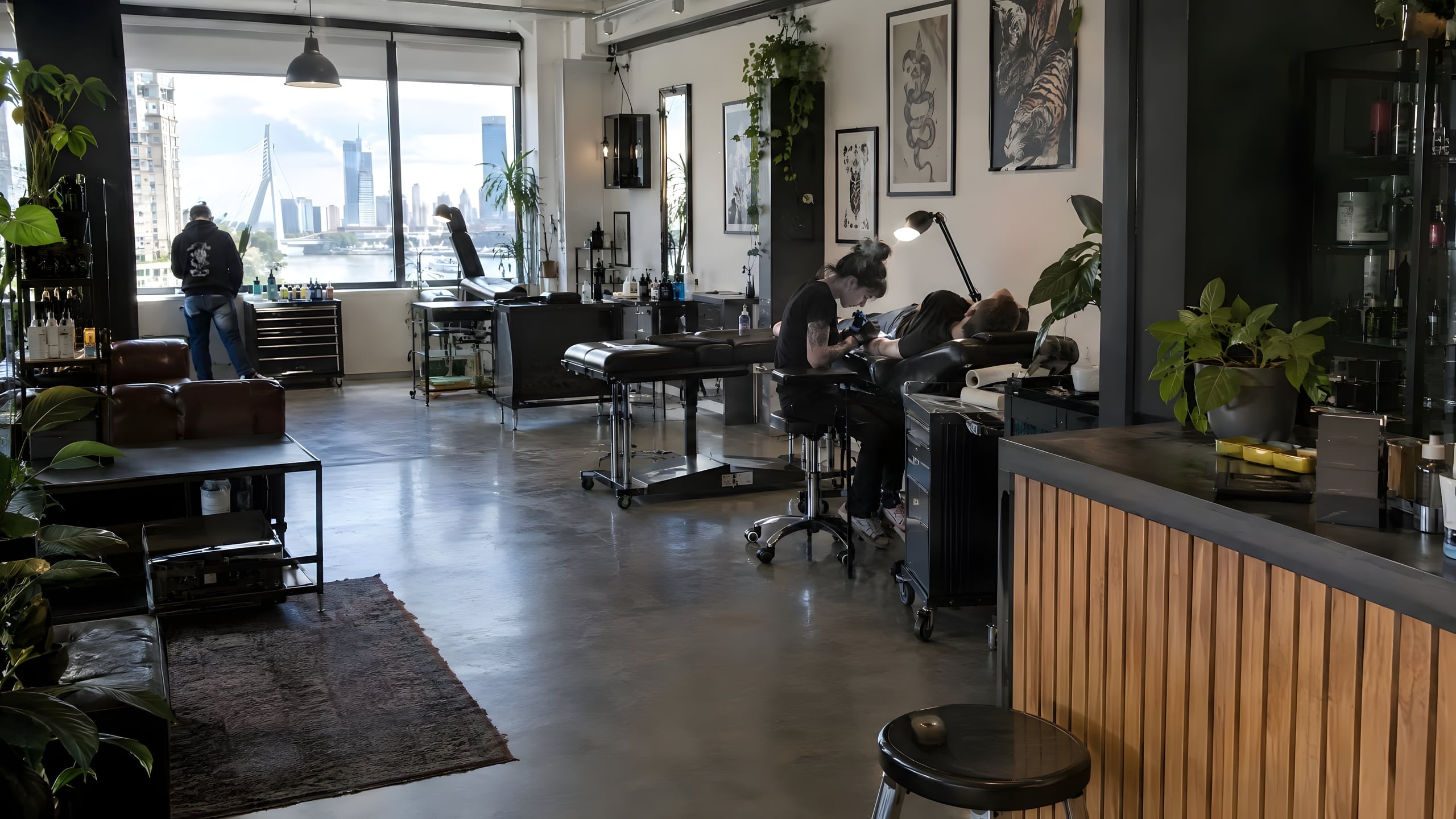 Tattoostudio met twee werkplekken, planten en hoge ramen met uitzicht over de rivier en de skyline.